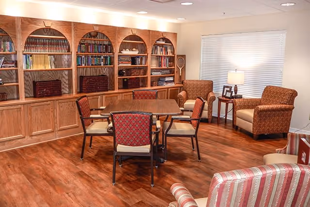 A cozy common area with wooden flooring, a wooden table surrounded by four chairs with patterned upholstery, two armchairs near a window with blinds, a side table with a lamp, and a large built-in wooden bookshelf filled with books and decorative items.