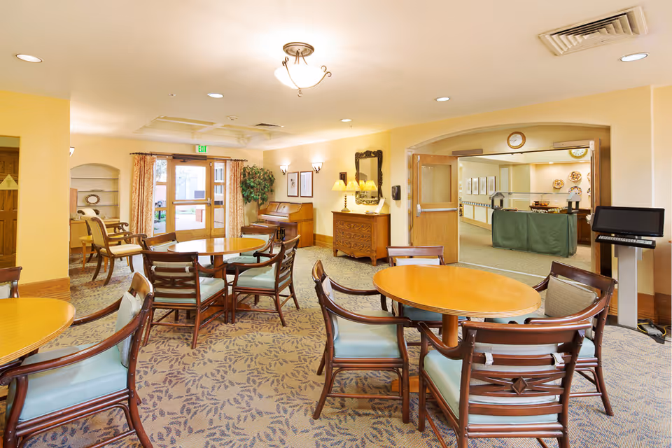 Bright communal dining room with round wooden tables and chairs, a piano and entry doors.