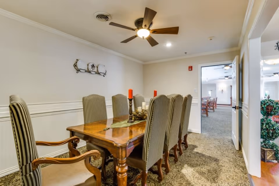 A formal dining room with a wooden table surrounded by eight high-backed striped chairs. The table is decorated with candles and a centerpiece. The room has beige walls with white wainscoting and a ceiling fan with a light fixture. An open door leads to another room with additional seating and decor.