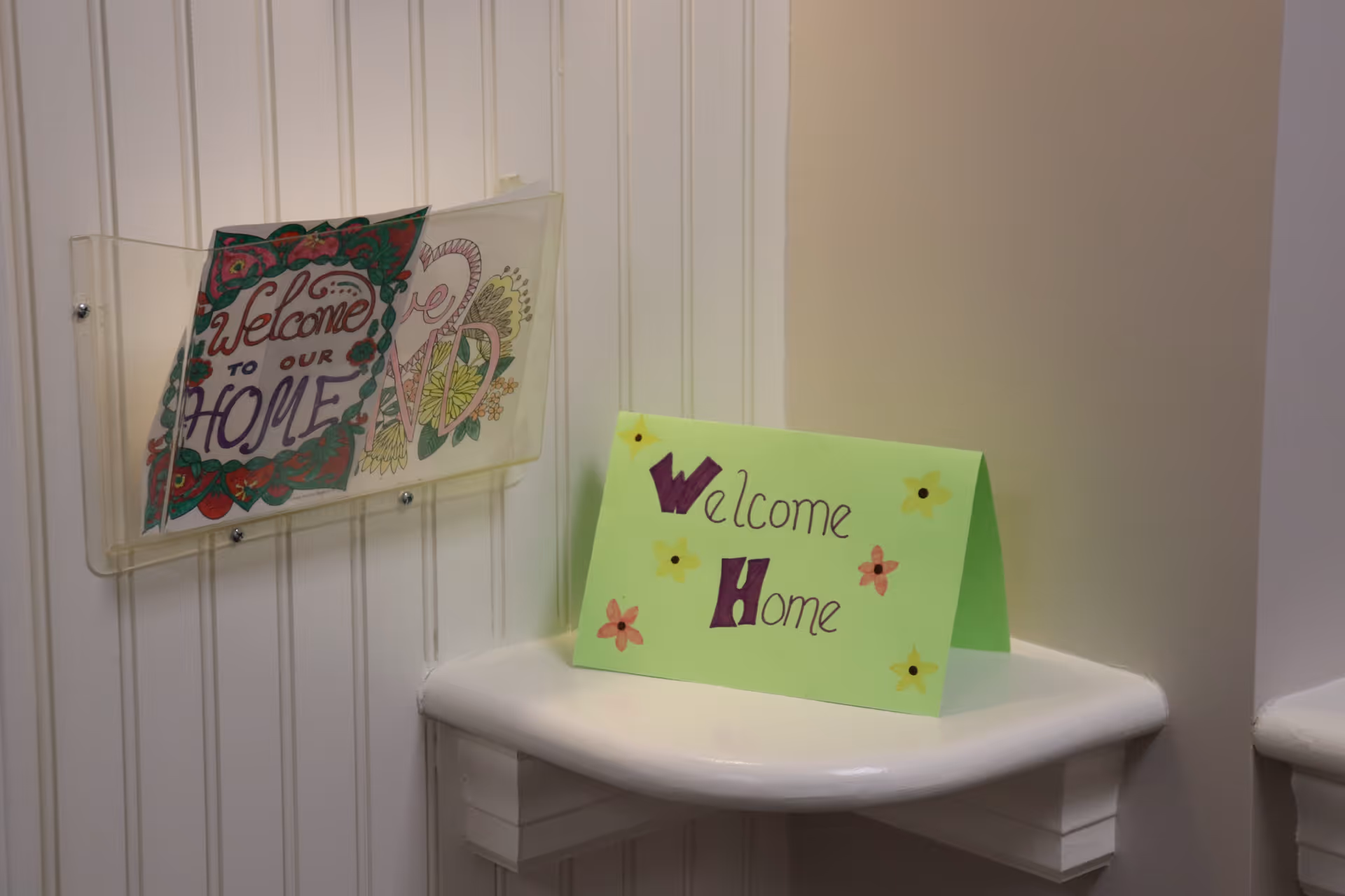 A small corner shelf with a green card that says 'Welcome Home' decorated with small flower drawings, next to a clear wall-mounted holder containing a colorful paper with the words 'Welcome to our Home' and decorative designs.