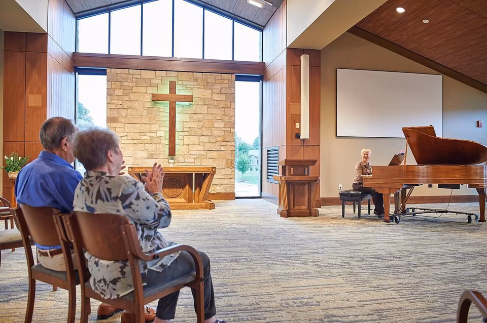 People seated in a chapel-style interior with a stone cross at the front and a woman playing a grand piano.