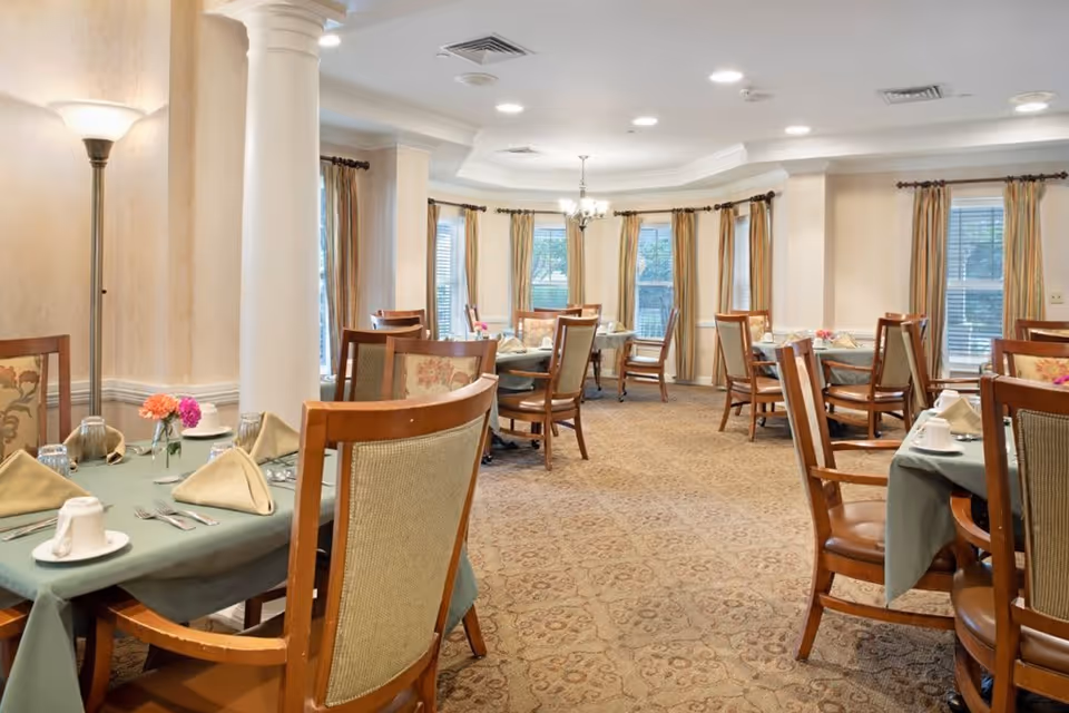 A well-lit dining room with multiple wooden tables and chairs arranged neatly. Each table is covered with a green tablecloth and set with folded beige napkins, white cups, glasses, and silverware. The room features large windows with beige curtains, a patterned carpet, and a white column near a floor lamp.