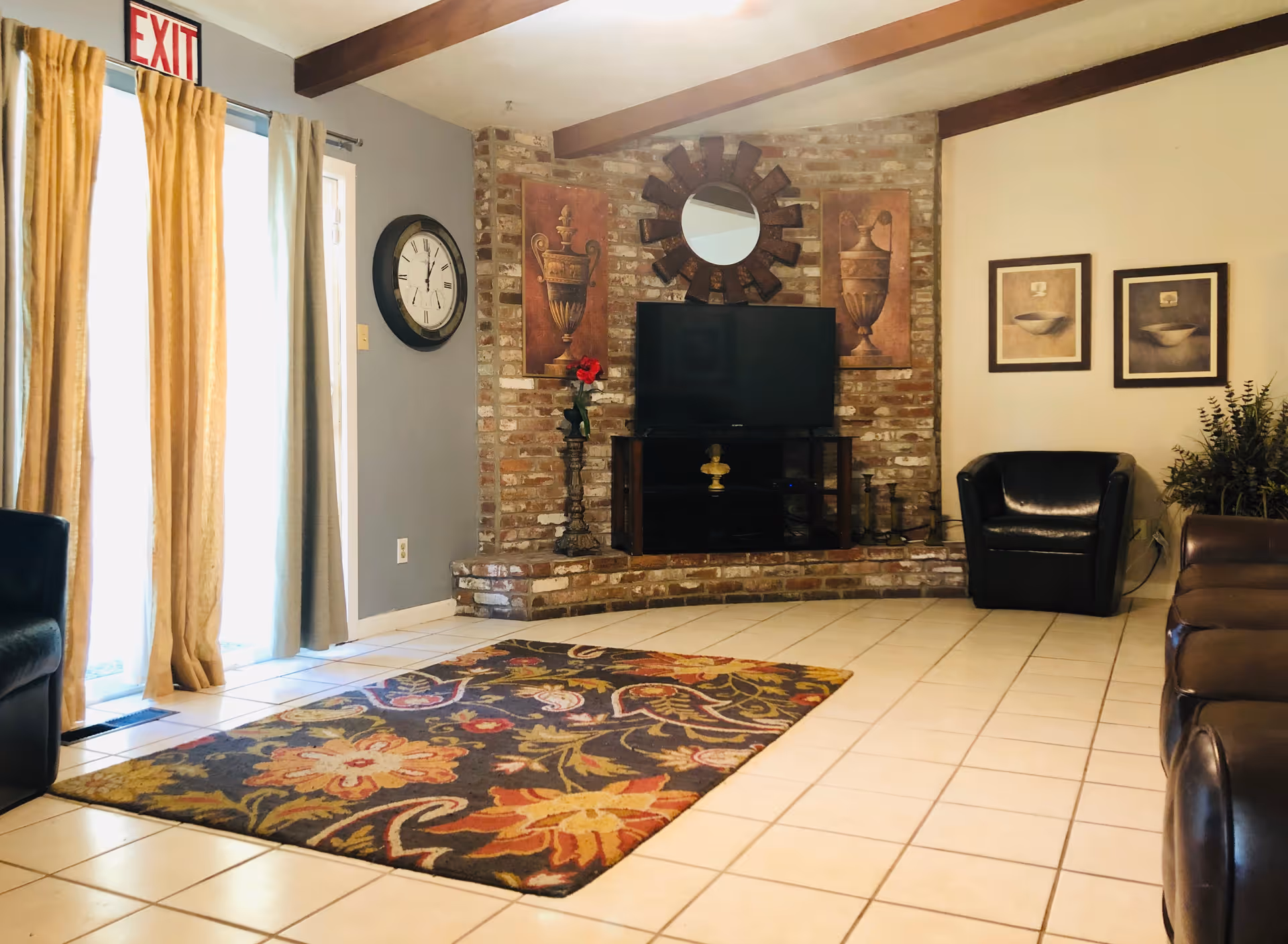A cozy living room with tiled floor and a colorful floral area rug. The room features a brick accent wall with a flat-screen TV on a stand, flanked by decorative artwork and a round mirror above. There are leather chairs and a sofa arranged around the room, with beige curtains covering a glass door and an exit sign above it. A large wall clock is mounted on a gray wall.