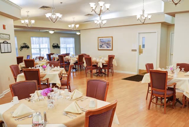 A well-lit dining room with multiple round tables covered in beige tablecloths, each set with folded napkins, glassware, and silverware. The room features wooden chairs with red upholstery, light wood flooring, cream-colored walls, and several ceiling light fixtures. There are windows with sheer curtains and some wall decorations including a framed picture and plants.