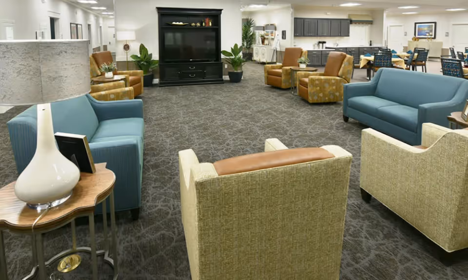 Spacious senior living common area with sofas and armchairs arranged around a large TV cabinet and dining tables in the background.