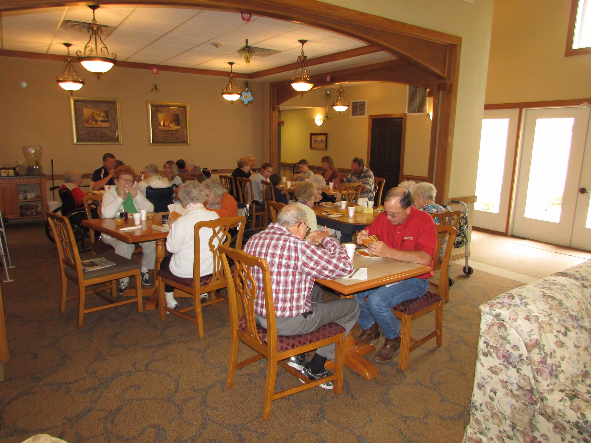 A dining room in a senior living facility with several elderly people and some younger individuals sitting at wooden tables eating and socializing. The room has carpeted floors, hanging light fixtures, framed artwork on the walls, and large windows letting in natural light.