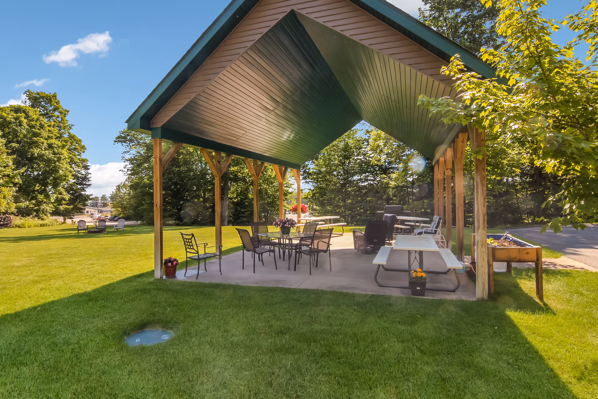 Outdoor covered pavilion with picnic tables, chairs, and flower pots on a concrete floor surrounded by green grass and trees under a blue sky with some clouds.