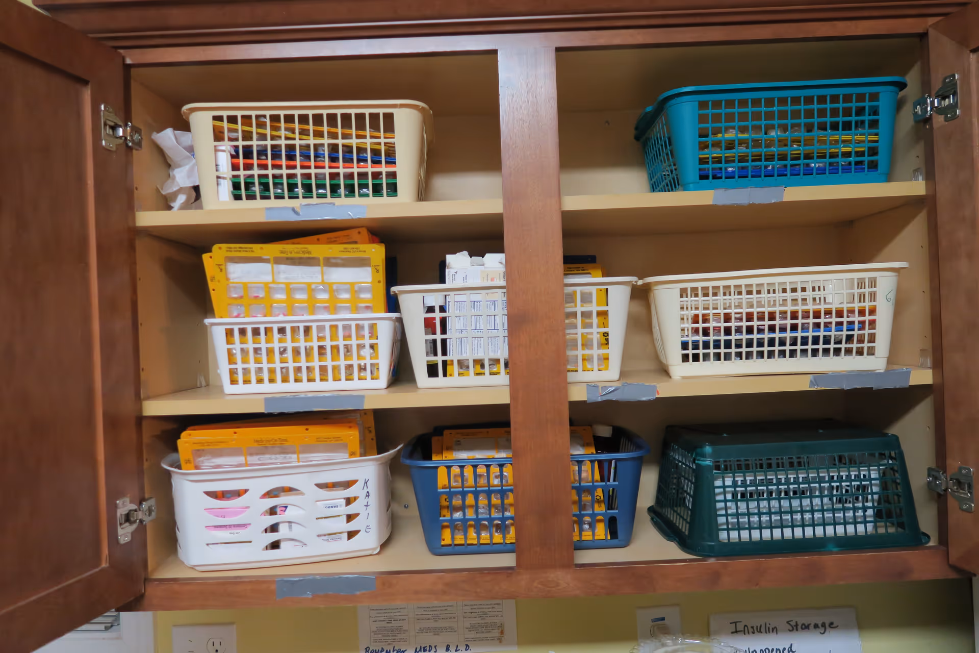 Open wooden cabinet shelves containing multiple plastic baskets filled with medication organizers and boxes, some labeled with names. Below the cabinet, a sign reads 'Insulin Storage'.