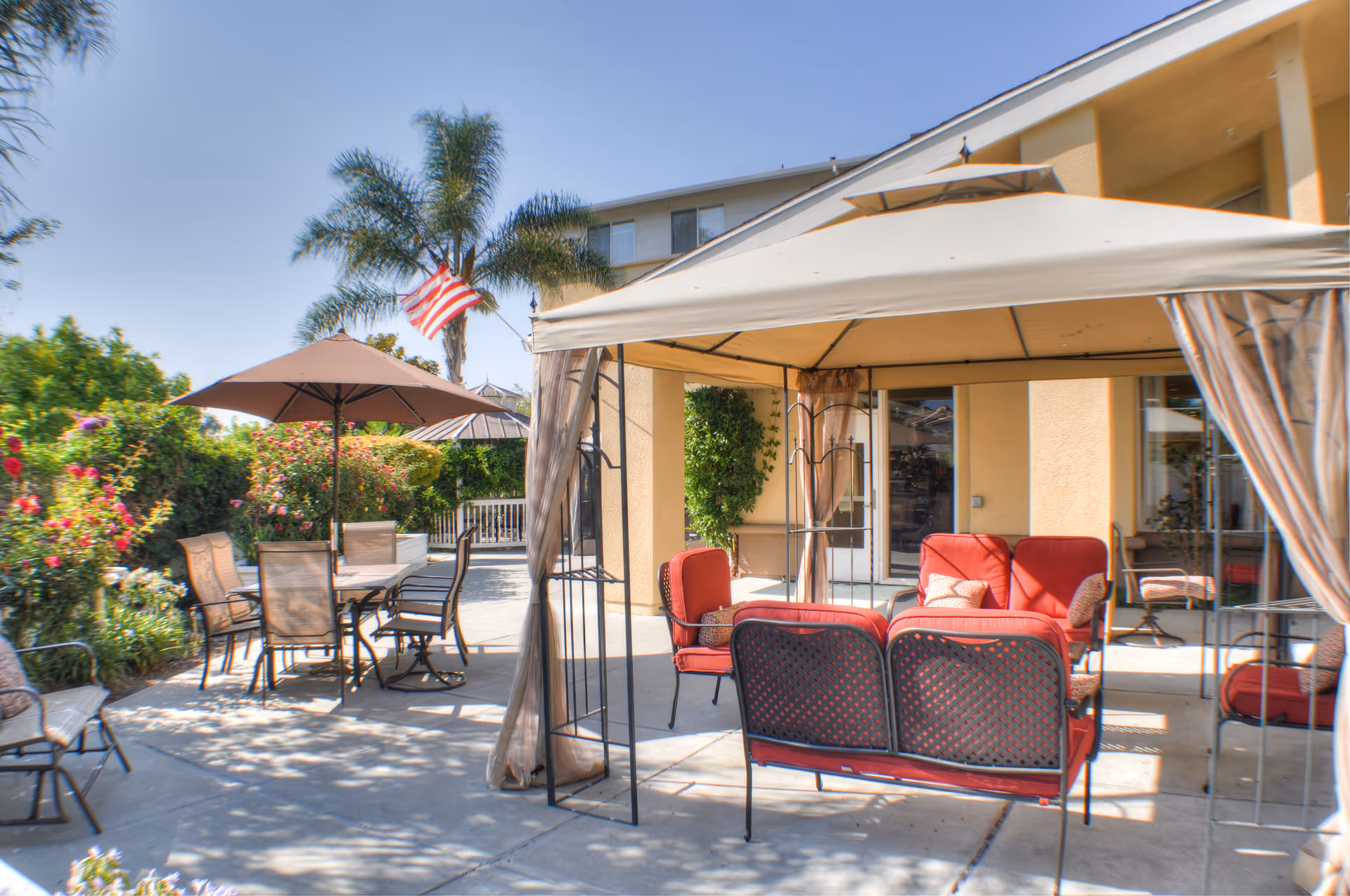 Outdoor patio area at Carlton Senior Living San Jose featuring a shaded seating area with red cushioned chairs and a loveseat under a canopy, a dining table with chairs and an umbrella, surrounded by greenery and palm trees under a clear blue sky.