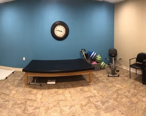 A therapy or exercise room with a large black padded treatment table in the center, a stationary exercise bike, several exercise balls, and a few chairs against the walls. The walls are painted blue and beige, and a round clock is mounted on the blue wall.