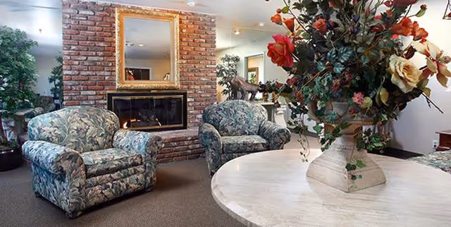 A cozy living room area featuring two floral upholstered armchairs facing a brick fireplace with a gold-framed mirror above it. A round white table with a large floral arrangement is in the foreground, and there are plants and decorative items around the room.