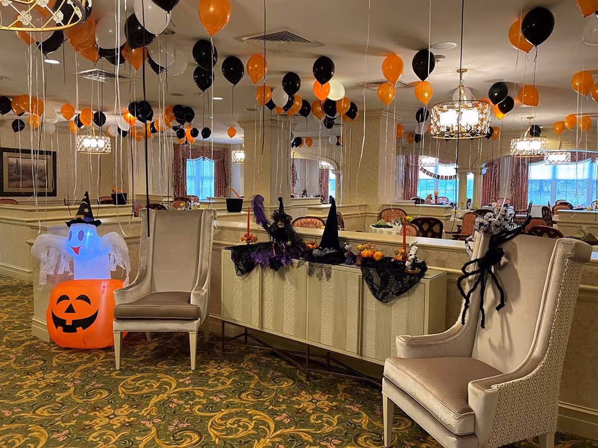 A decorated interior room with Halloween-themed decorations including black and orange balloons hanging from the ceiling, a glowing ghost figure with a witch hat, a large pumpkin decoration, witch hats, candles, and a large spider on a beige armchair. The room has patterned carpet, mirrors on the walls, and several chandeliers.