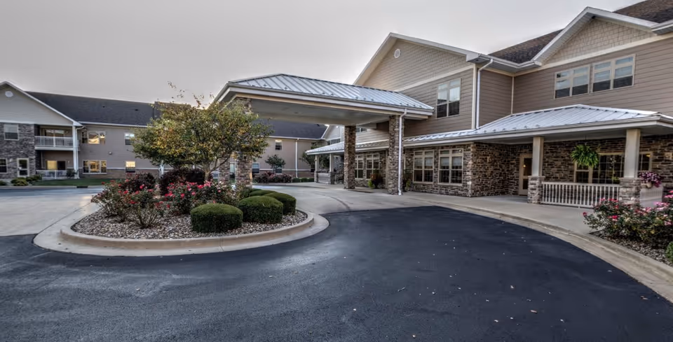 Exterior view of Lincolnshire Place of Decatur showing the entrance with a covered drop-off area, landscaped bushes and flowers, and a multi-story building with stone and siding exterior.