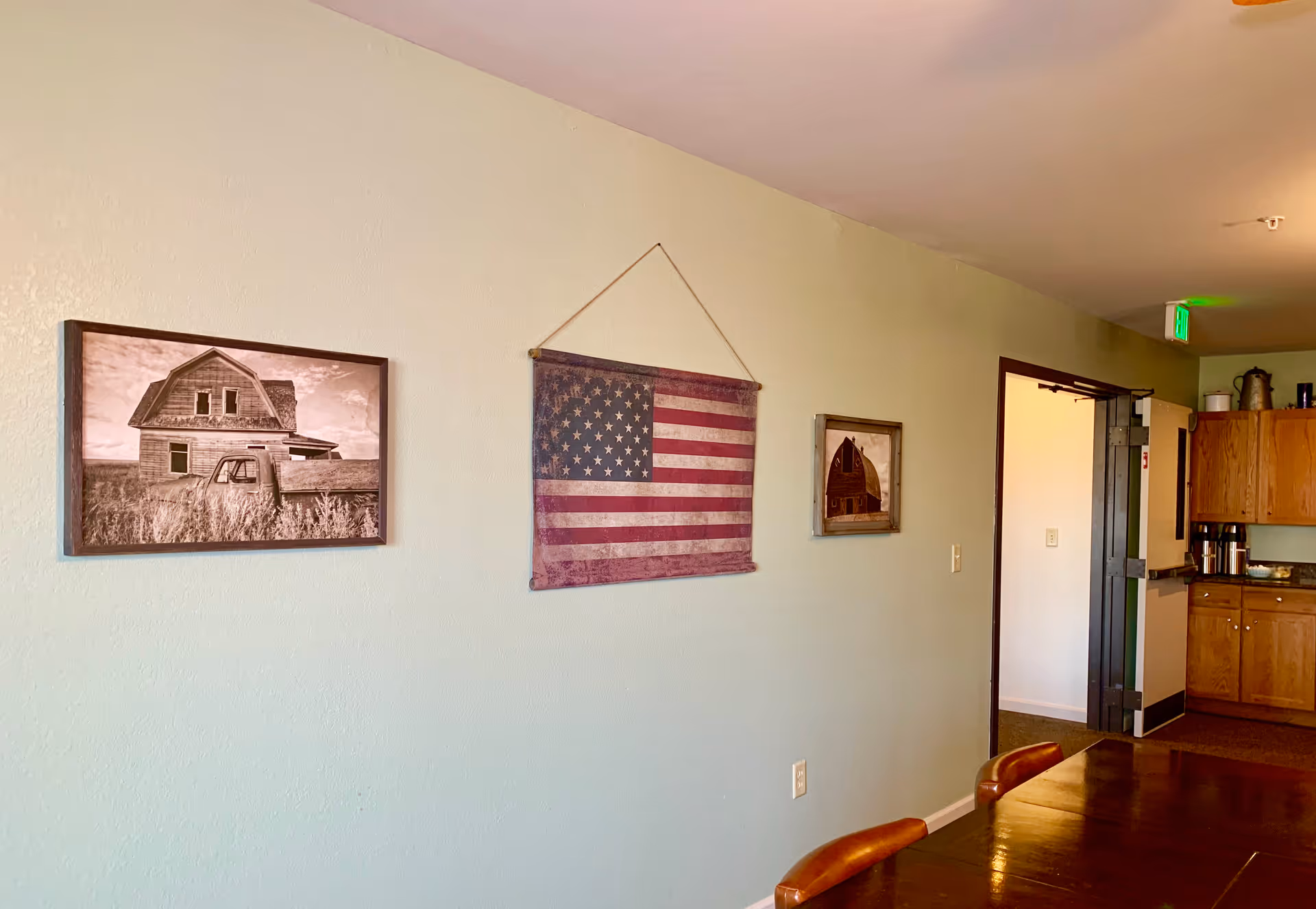 Interior communal dining area with a wooden table, wall decorations including a hanging American flag and framed photos, and an open doorway to a kitchenette.