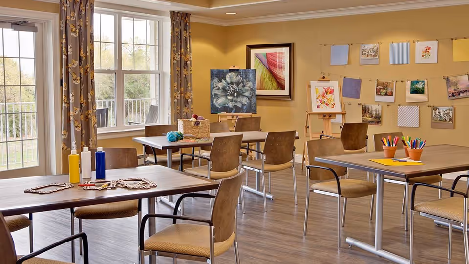 A bright activity room with several tables and chairs arranged for arts and crafts. The tables hold paint bottles, colored pencils, and craft supplies. The walls are decorated with framed artwork and various pictures clipped to strings. Large windows with floral curtains let in natural light, and a door leads outside to a balcony or patio area.