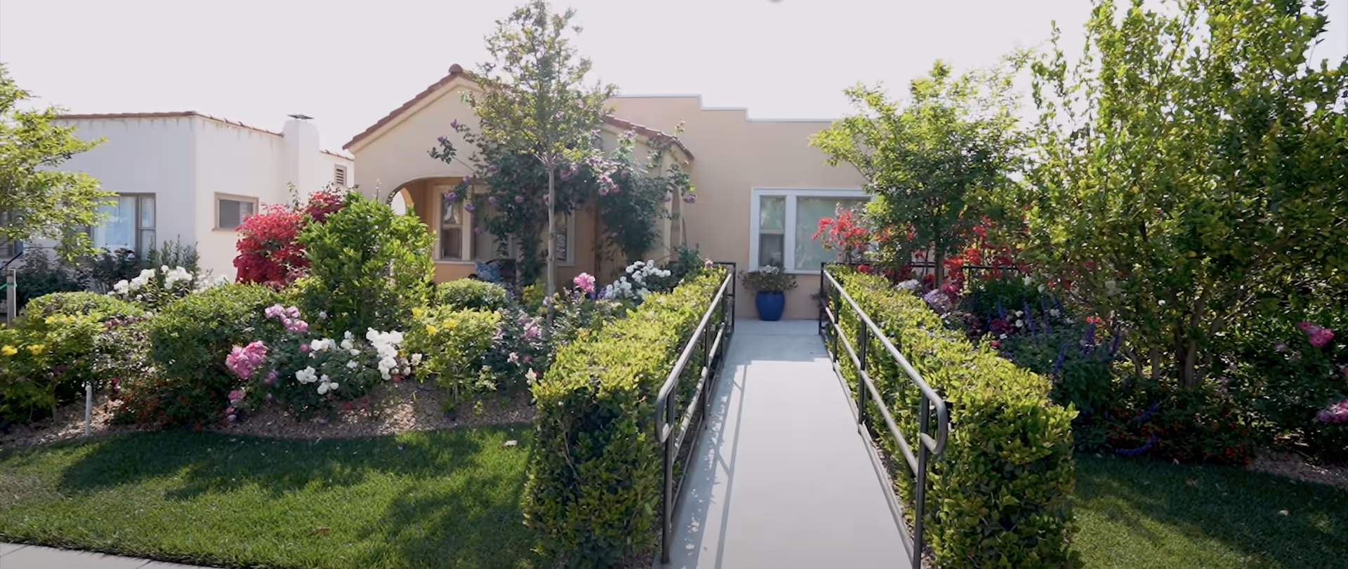 Front exterior view of a single-story building with a tiled roof and a ramp leading to the entrance, surrounded by lush green bushes, trees, and colorful flowers.