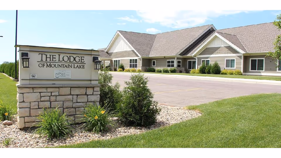 Stone sign reading "The Lodge of Mountain Lake" in front of a single-story residential building with landscaped lawn and parking lot.