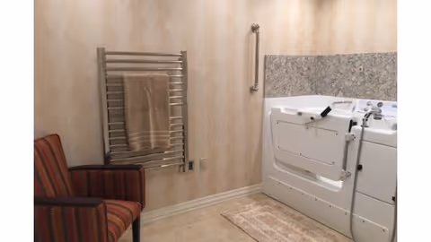 Bathroom with a walk-in tub, wall-mounted grab bar, towel warmer, small rug, and a striped armchair.