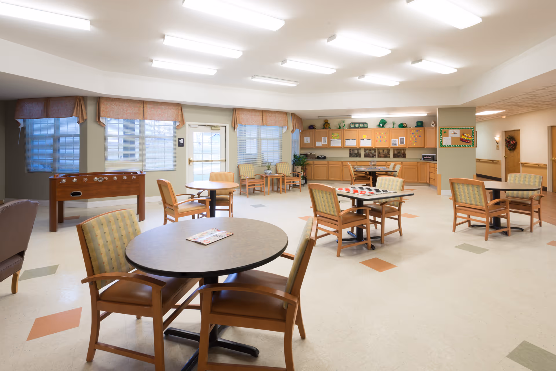A spacious common area with multiple round tables and chairs arranged for seating. The room features large windows with valances, a foosball table, and a game table with checkers. There are cabinets and a countertop along the back wall with various decorations and plants. The floor has a pattern of colored tiles, and the ceiling is fitted with bright fluorescent lights.