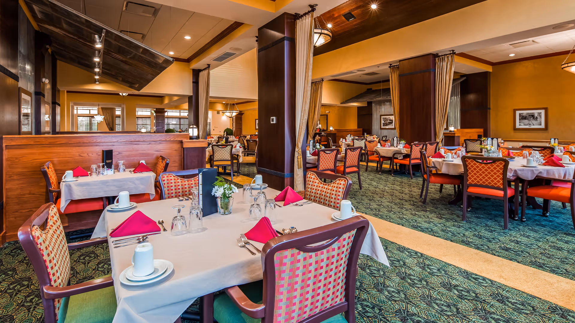 A spacious dining room in a senior living facility with multiple tables set with white tablecloths, red folded napkins, cups, glasses, and silverware. The room features patterned carpet, wooden pillars, warm yellow walls, and large windows allowing natural light. Chairs have a red and beige checkered pattern, and there are framed pictures on the walls.