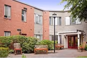 Two-story brick and wood-clad facility courtyard with benches, landscaping, a lamppost, and a covered entrance.