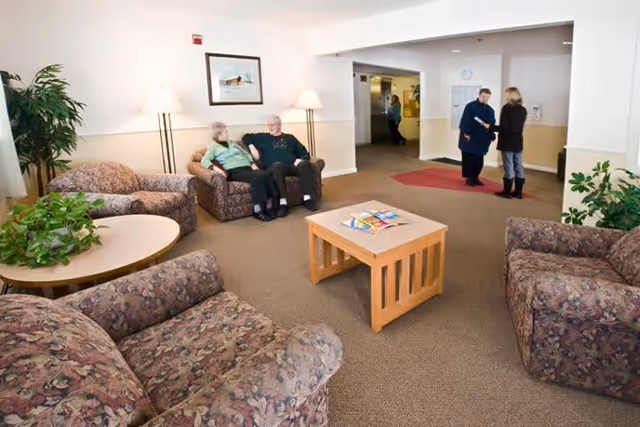 A senior living facility common room with floral-upholstered sofas, a wooden coffee table, plants, and residents chatting.
