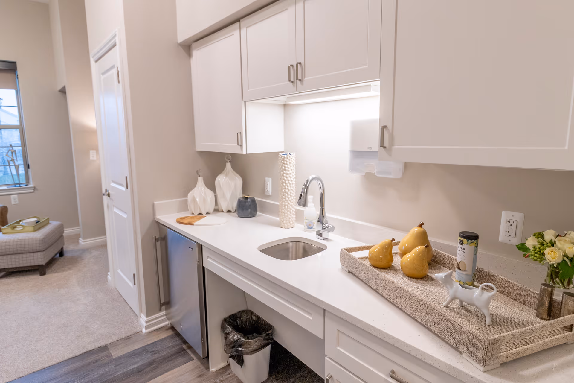 Small modern kitchenette with white cabinets, a sink, countertop decor including a tray with yellow ceramic pears, and a mini fridge opening to a carpeted living area.