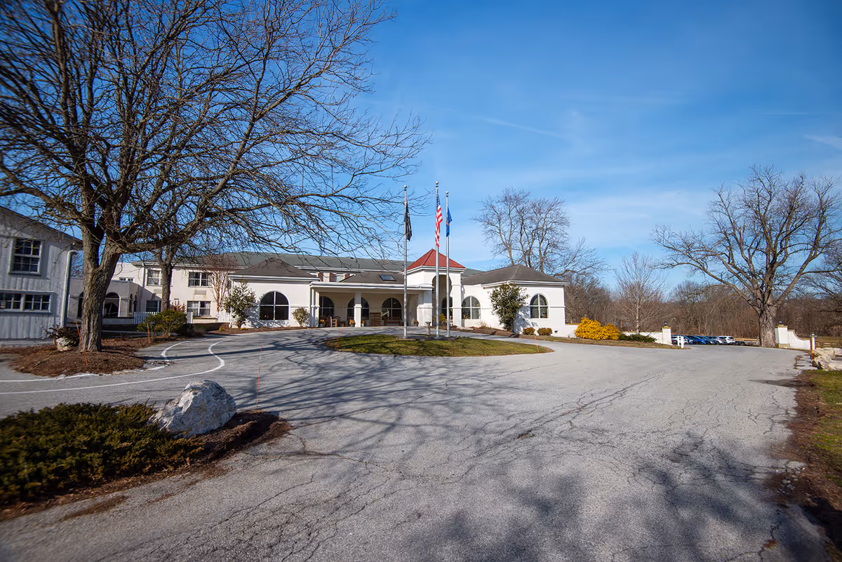 Front exterior of a nursing and rehabilitation center with a circular driveway, flagpoles, and the main entrance under a clear blue sky.