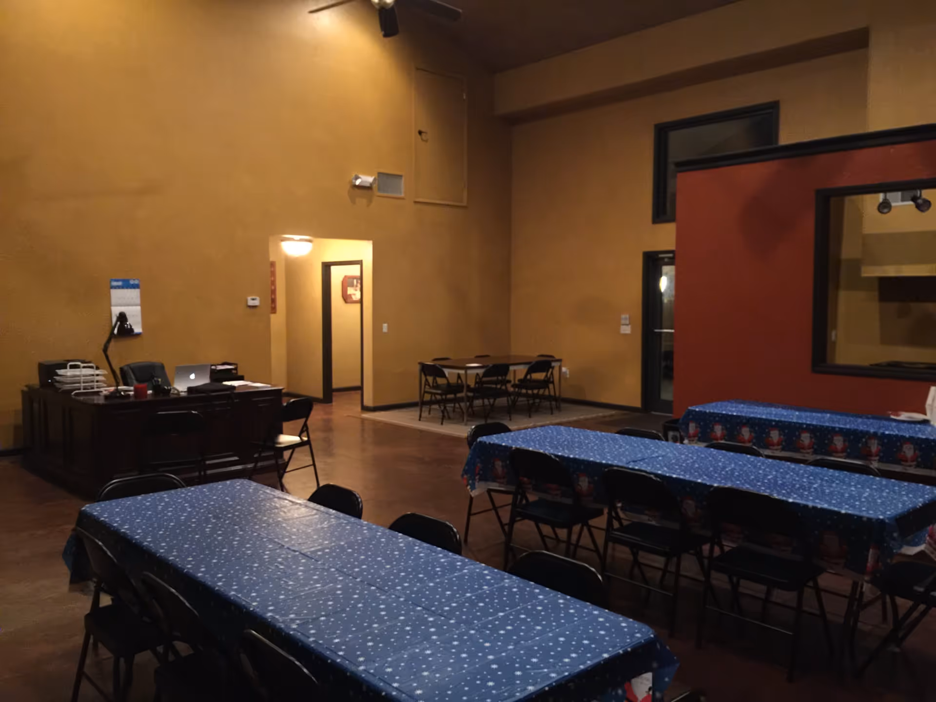 Large multipurpose dining room with long tables covered in blue tablecloths, folding chairs, and a desk against a high tan wall.