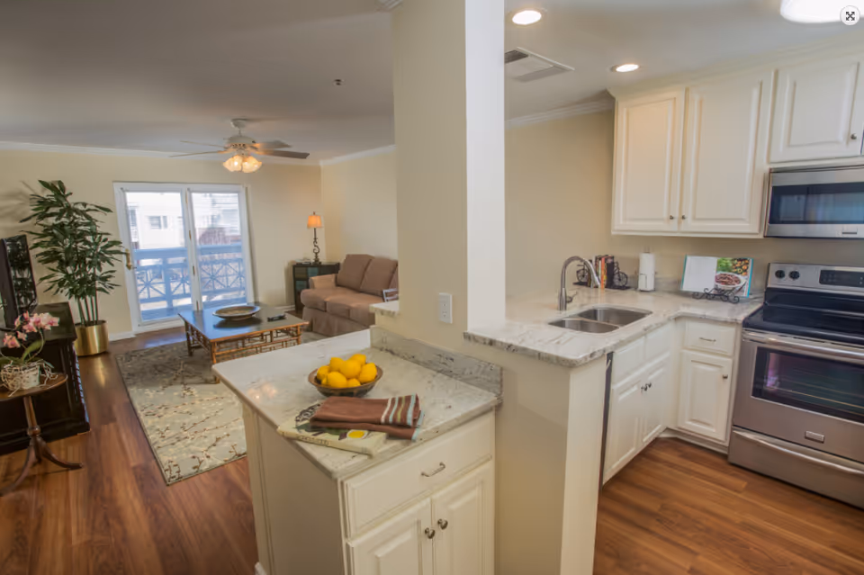 Open-plan apartment interior showing a bright kitchen with marble countertops opening to a living room with a sofa and sliding glass doors.