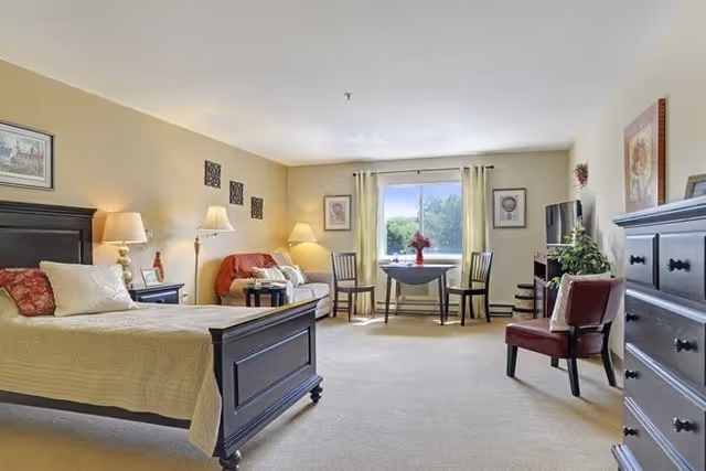 A spacious senior living bedroom with a large bed, two bedside tables with lamps, a small sofa with a throw blanket, a round table with two chairs near a window with curtains, a TV on a stand, a red armchair, and a dresser. The room is well-lit with natural light coming through the window and decorated with framed artwork on the walls.