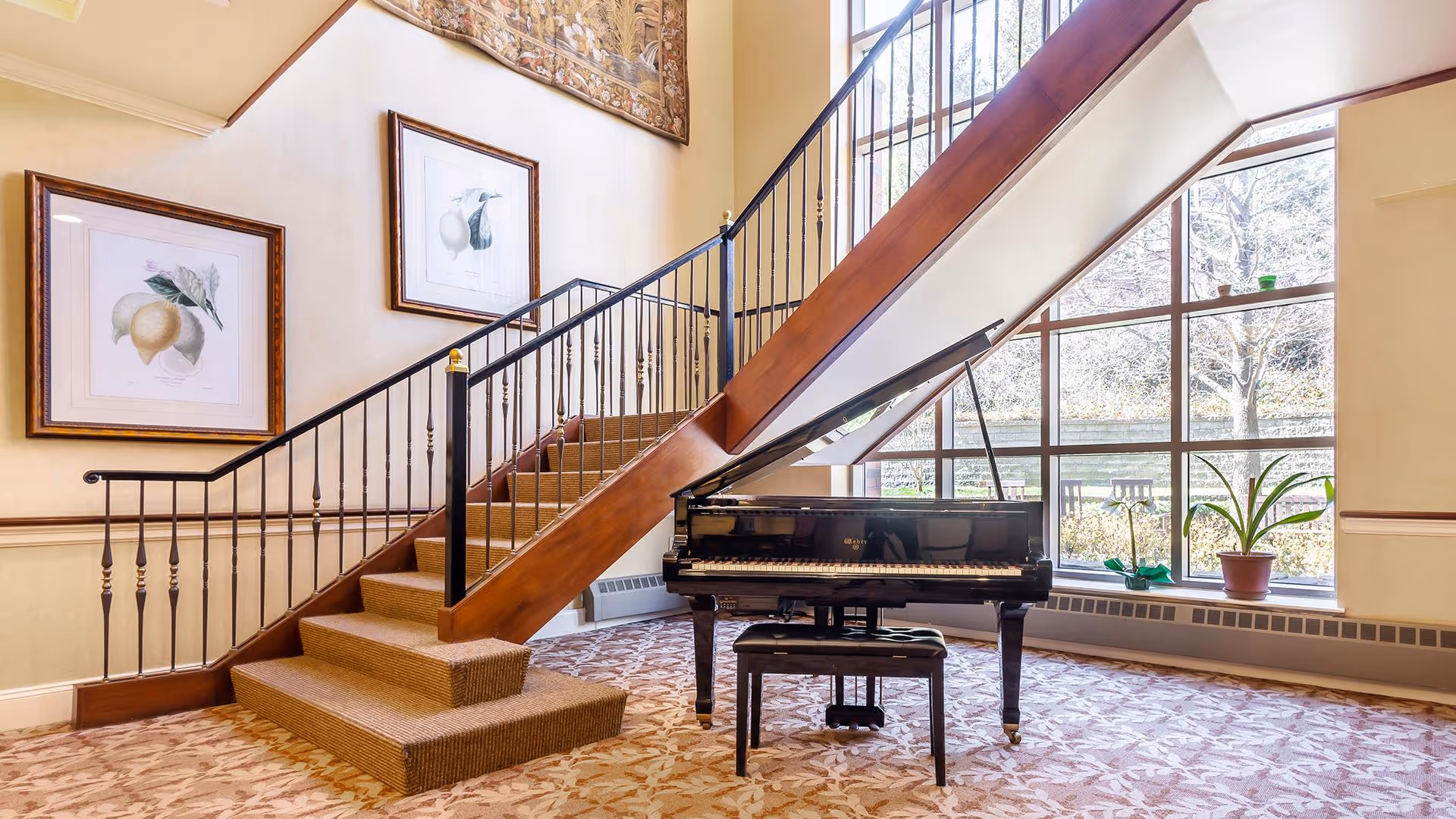 Interior view of a senior living facility showing a grand piano with an open lid and a piano bench in front of a large window. To the left, there is a carpeted staircase with a wooden handrail and black metal balusters. The walls are decorated with framed botanical prints and a tapestry above the staircase. The floor is covered with a patterned carpet.