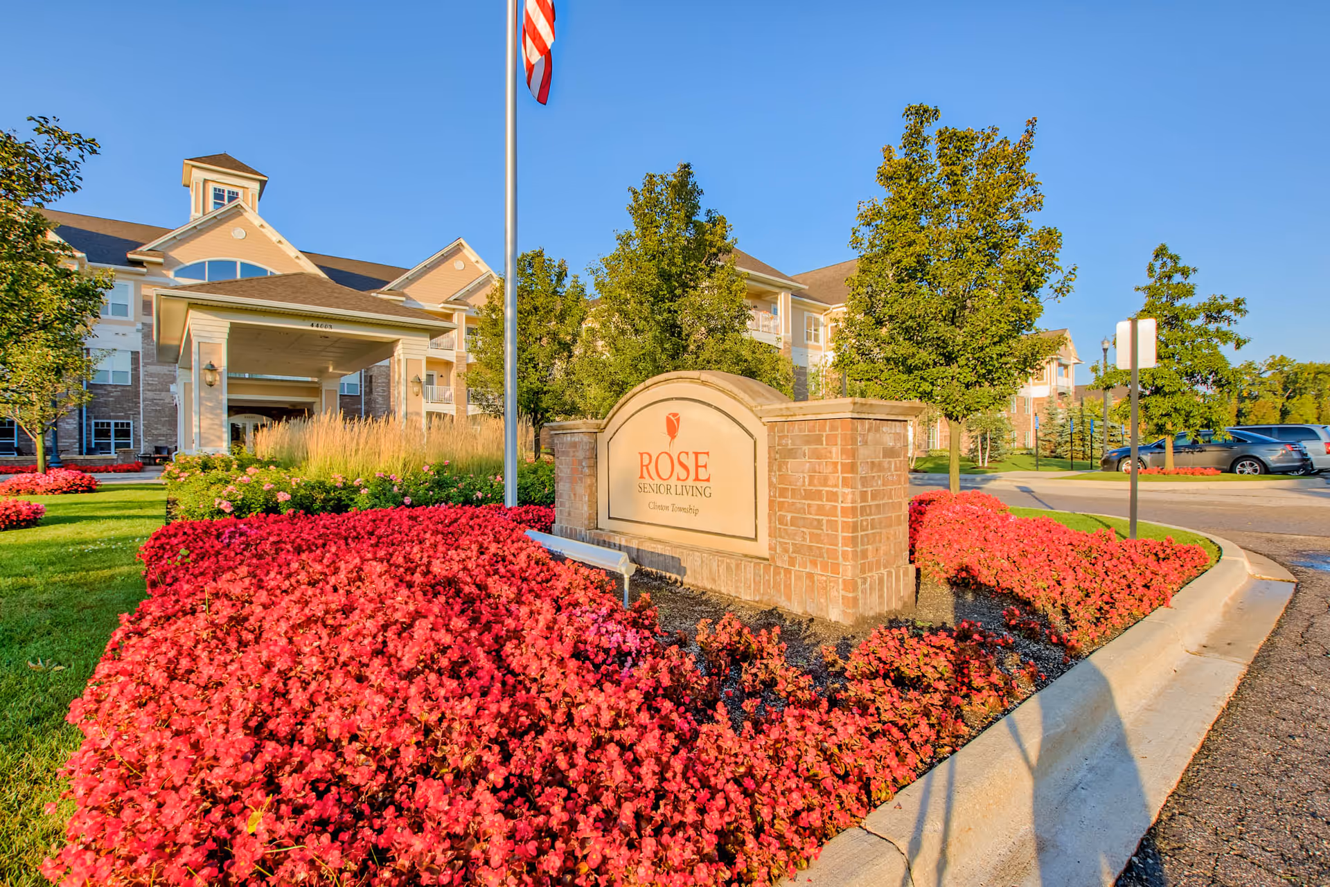 Entrance to Rose Senior Living Clinton Township featuring a brick sign with the facility name, surrounded by vibrant red flowers and green trees, with the building and a flagpole in the background under a clear blue sky.