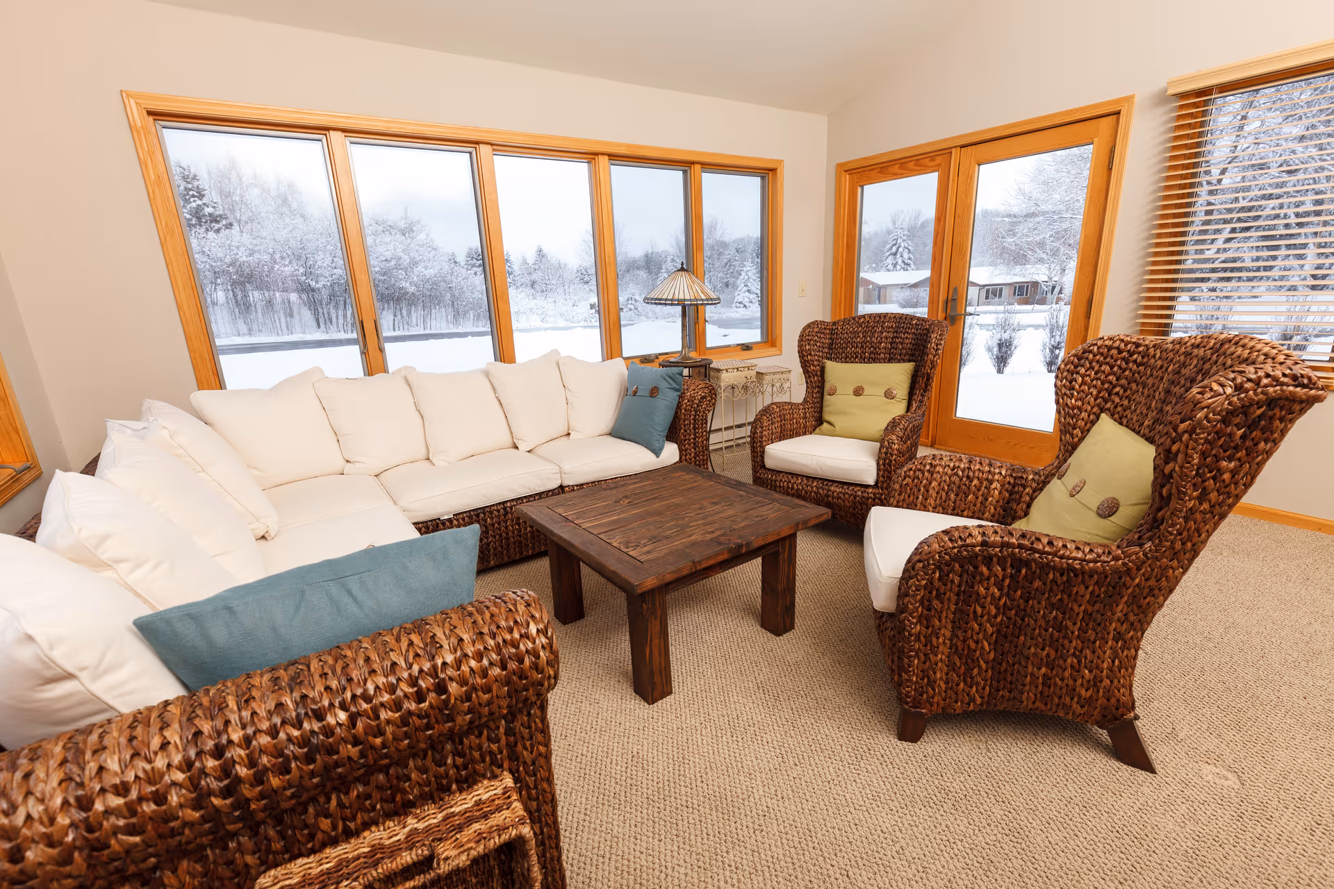 Cozy living room with a white sectional, woven armchairs and a wooden coffee table in front of large windows showing a snowy outdoor scene.