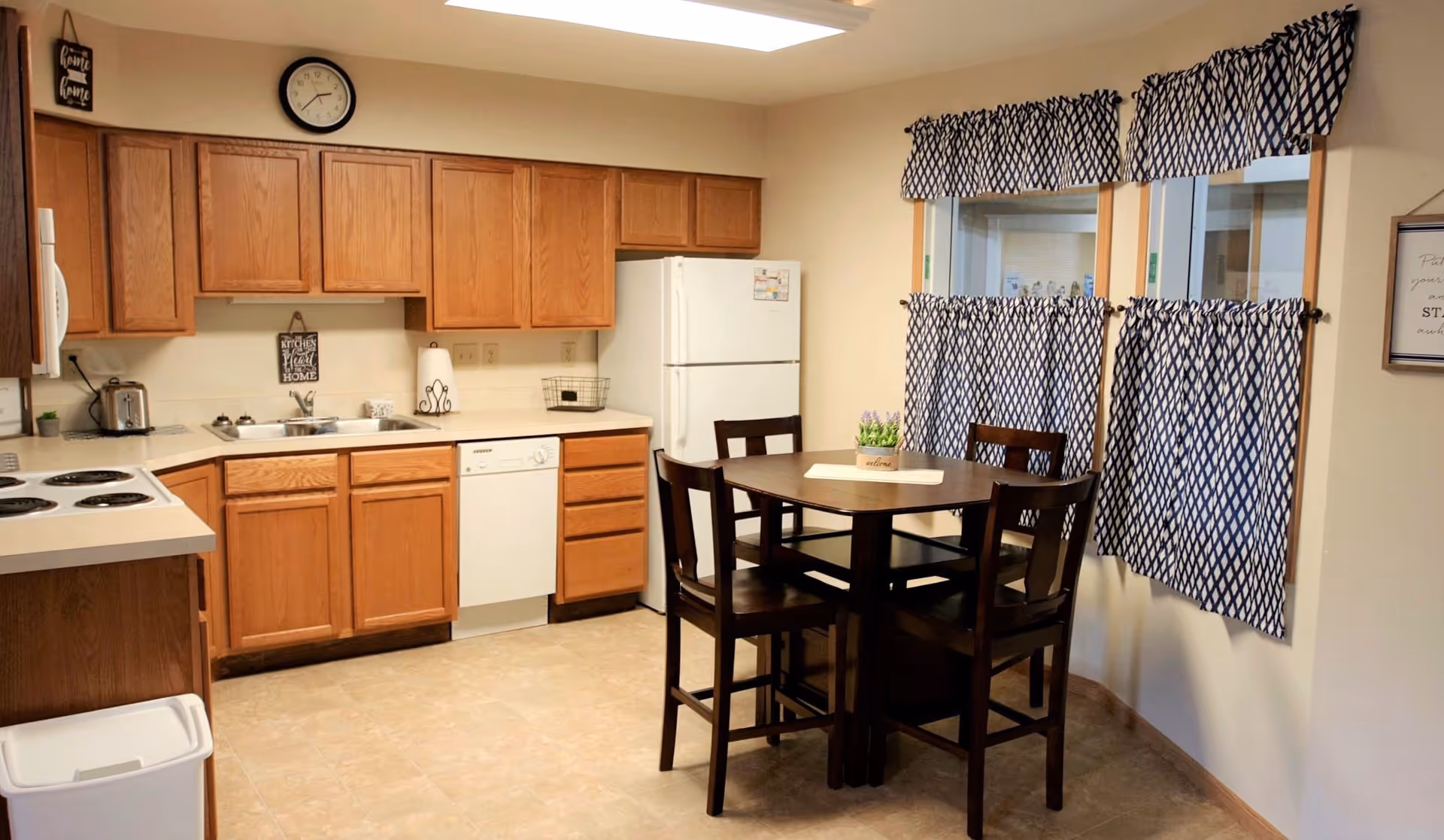 Open kitchen with oak cabinets, white appliances, and a small dark-wood dining table with four chairs by windows with patterned curtains.