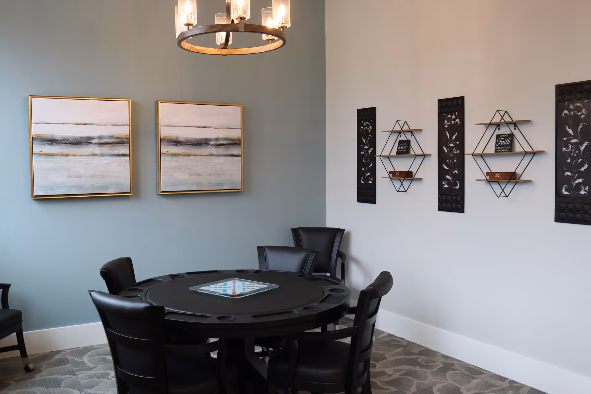 A small room with a round black table and five black chairs around it. The table has a board game set up in the center. The walls are painted in two colors, light blue and white, and decorated with two abstract paintings and three black decorative wall hangings with shelves holding small items. A modern chandelier hangs from the ceiling.