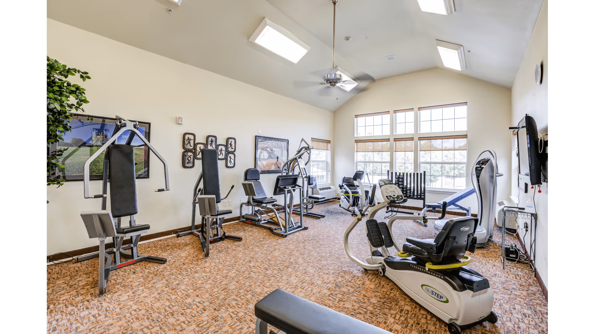 A bright fitness room with various exercise machines including weight machines, a recumbent bike, and other cardio equipment. The room has large windows letting in natural light, a ceiling fan, and light-colored walls with framed pictures and a plant in the corner.