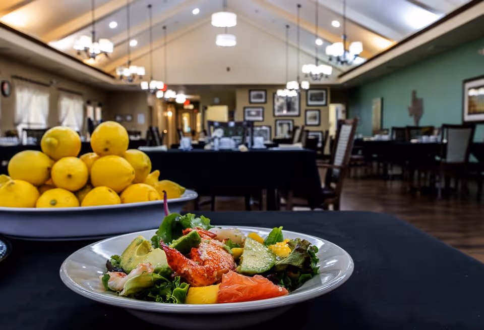 A close-up of a fresh salad with avocado, leafy greens, grapefruit, and lobster on a white plate on a black tablecloth. In the background, there is a large bowl filled with lemons and a spacious dining room with tables, chairs, chandeliers, and framed pictures on the walls.