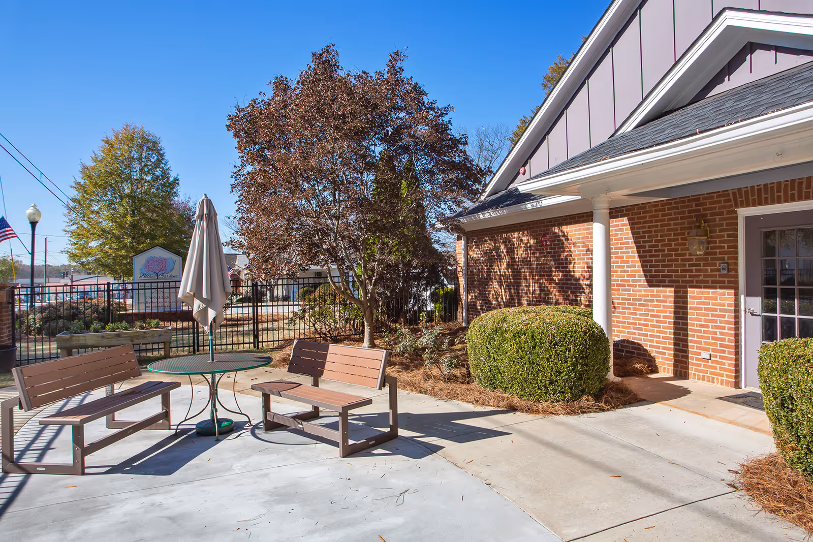 Outdoor patio area at Bethel Gardens Assisted & Memory Care featuring two wooden benches and a round table with a closed umbrella. The patio is surrounded by bushes, a tree with brown leaves, and a brick building with a white column and a glass door. A black metal fence and a sign are visible in the background under a clear blue sky.