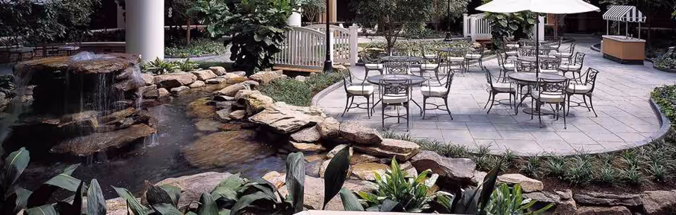 Outdoor patio area with several metal tables and chairs, some shaded by large white umbrellas. In the foreground, there is a small rock waterfall flowing into a pond surrounded by lush green plants. The area is paved with stone tiles and bordered by greenery and trees.