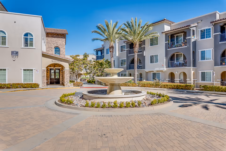 Exterior view of Meridian at Lake San Marcos senior living facility featuring a circular driveway with a central stone fountain surrounded by small plants and palm trees, with multi-story residential buildings in the background under a clear blue sky.