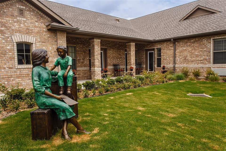 A brick senior living building courtyard with a lawn and a bronze statue of a woman reading to a child.