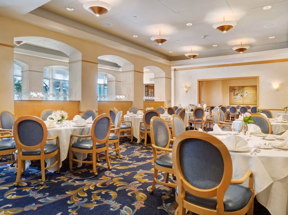 Bright, elegant dining room with round white-clothed tables and wooden chairs set for a meal.