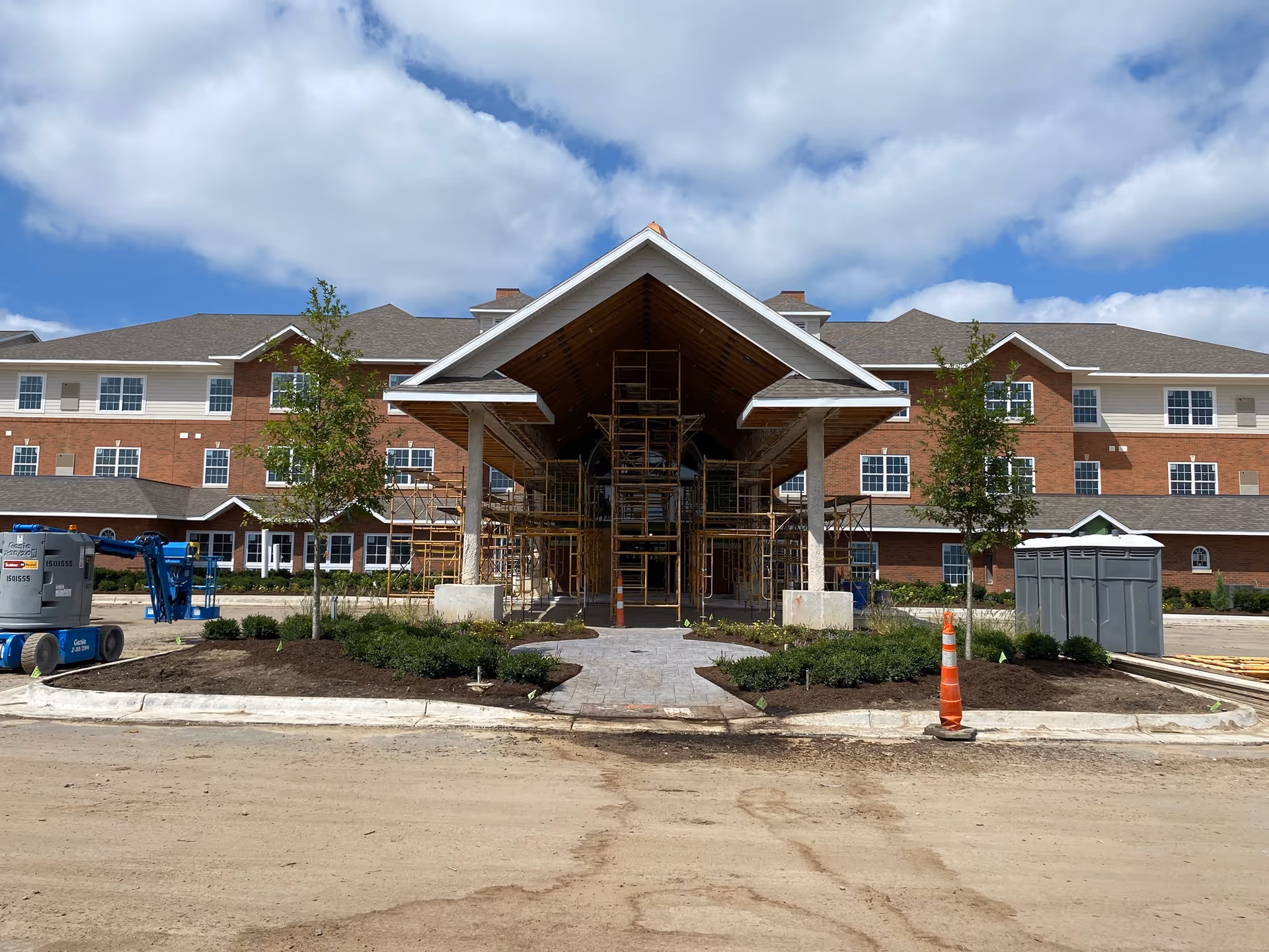 Front exterior view of a large brick and siding building under construction with scaffolding around the entrance canopy, newly planted landscaping, a construction lift on the left, and a portable toilet on the right under a partly cloudy sky.