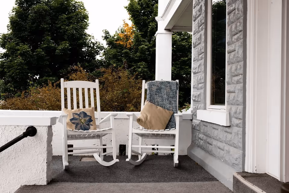 Two white wooden rocking chairs on a porch with cushions and a blanket, next to a stone wall and window, with green trees and bushes in the background.