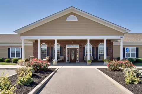 Front exterior view of a single-story senior living facility building with a covered entrance supported by white columns, brick and beige siding, landscaped bushes and flowers along the driveway, and a clear blue sky.