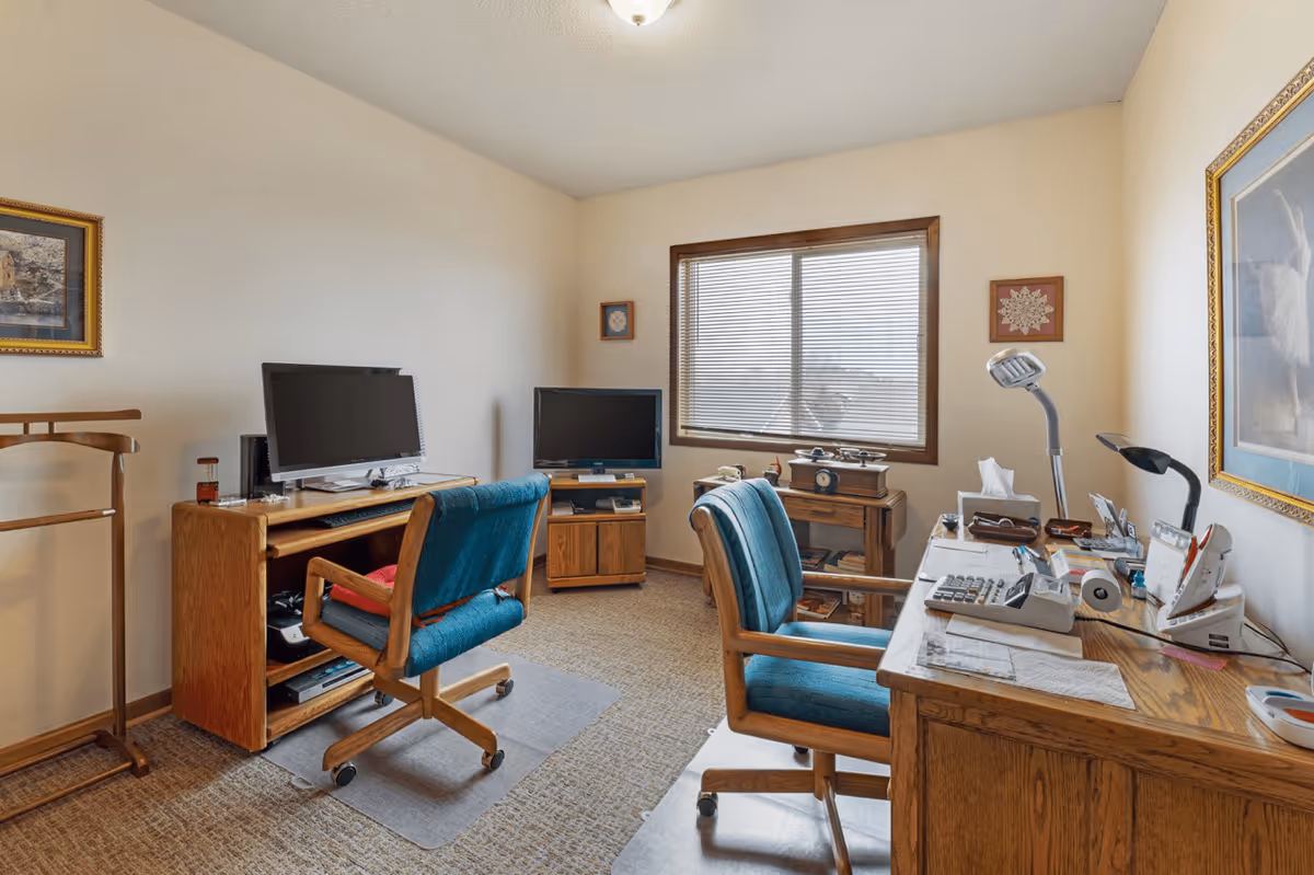 A small office or study room with two wooden desks, two teal upholstered office chairs on wheels, a computer monitor, a television, a desk lamp, and various office supplies. The room has beige walls, a window with blinds, and framed artwork on the walls.