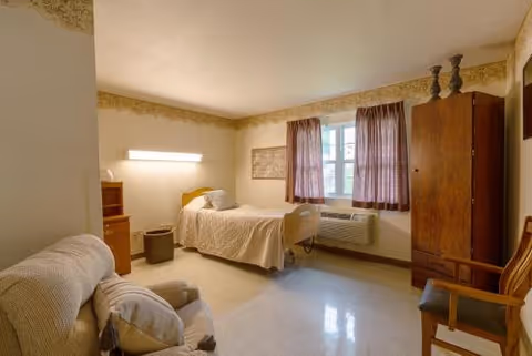 A simple skilled nursing bedroom with a single bed, armchair, wooden wardrobe, and a window with curtains.
