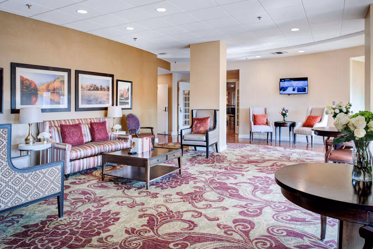 A spacious and well-lit living room area with patterned carpet in shades of red and beige. The room features a striped sofa with red cushions, several armchairs, a wooden coffee table, side tables with lamps, and framed landscape artwork on the wall. A flat-screen TV is mounted on the far wall, and there are flower arrangements on tables throughout the room.