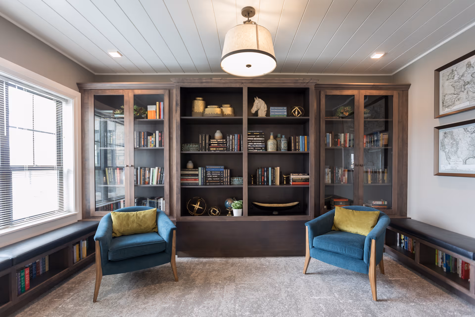 A cozy reading room with two blue armchairs each with a yellow cushion, positioned in front of a large dark wooden bookshelf filled with books and decorative items. The room has a window with blinds on the left, built-in bench seating with books underneath on both sides, a light fixture hanging from the white paneled ceiling, and framed maps on the right wall.